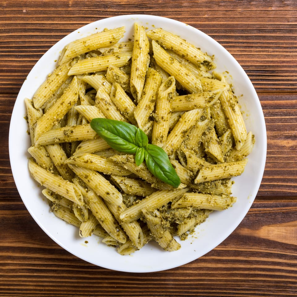 Penne pasta with vibrant green pesto sauce and a fresh basil leaf on a white plate.